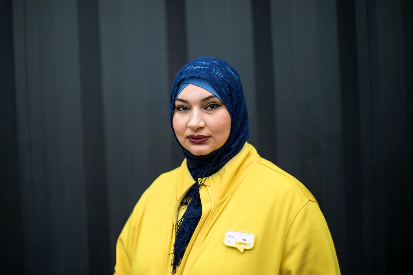 Uchodźczyni -Aza. Uśmiechnięta kobieta w żółtej bluzie z granatową chustą na głowie patrzy w kamerę.