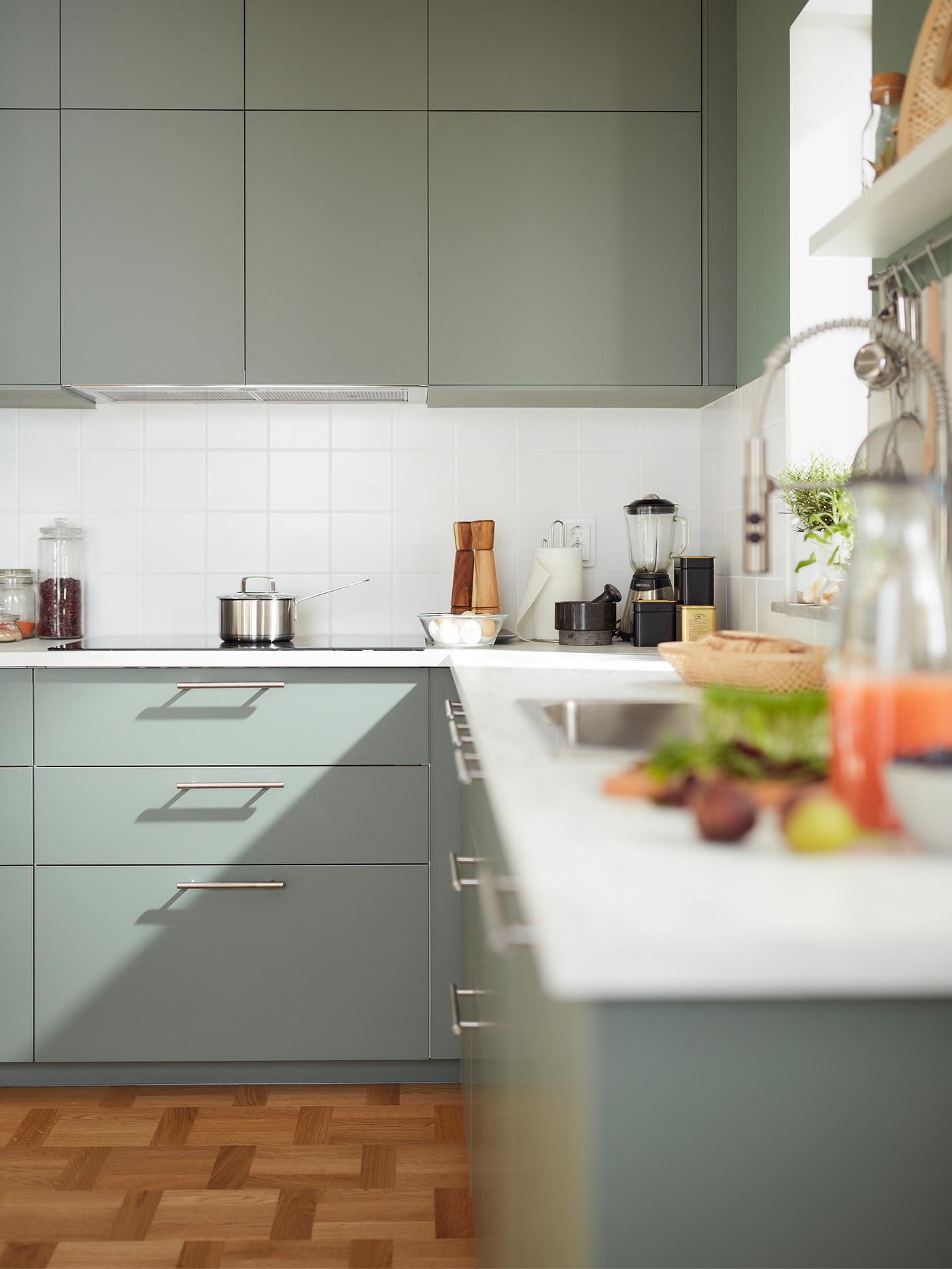 Ugaona kuhinja sa sivozelenim ormarićima, grejnom pločom i svetlosivom radnom pločom s imitacijom minerala, s priborom i uređajima na vrhu. 