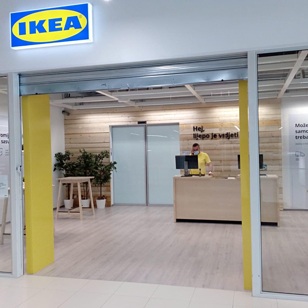 Ulaz u IKEA dostavni centar s IKEA logotipom iznad ulaza, drvenim zidovima i radnikom za pultom. Scena prikazuje ulazni prostor trgovine s karakterističnim IKEA dizajnom.