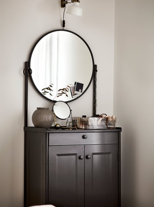 Um armário KORNSJÖ em preto com um grande espelho redondo, com objetos decorativos expostos, por cima do qual está um candeeiro branco.