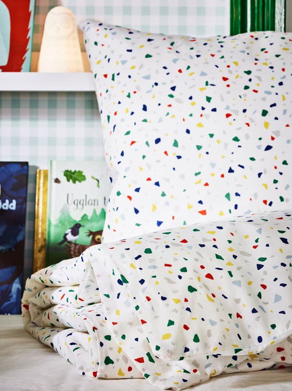 Um conjunto de roupa de cama MÖJLIGHET numa cama por fazer, com um candeeiro LED SOLBO e livros em prateleiras para molduras na parede atrás.