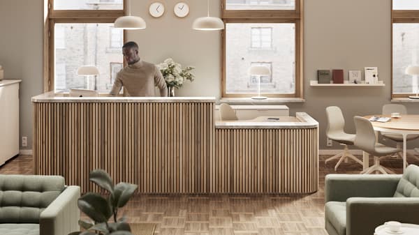 Um homem atrás de uma secretária de receção em madeira clara, à frente de duas janelas. Dois candeeiros suspensos pendurados em cima e relógios na parede.