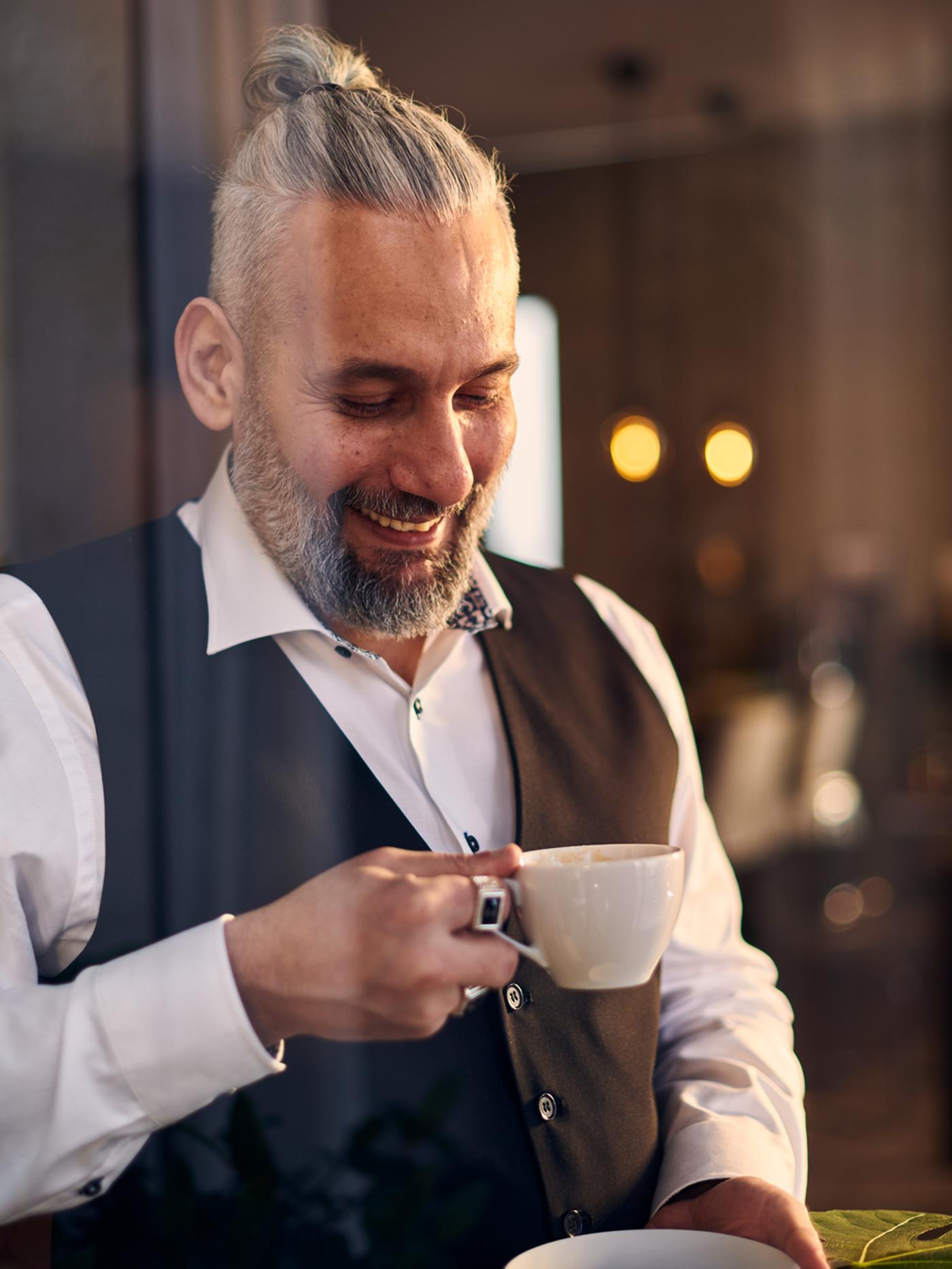 Um homem de colete e camisa branca a sorrir, com um pires numa mão e uma chávena na outra.