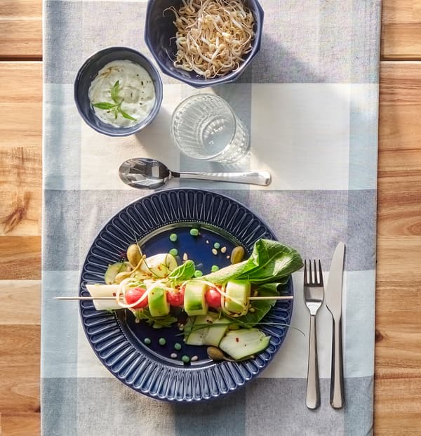 Um prato azul escuro com vegetais coloridos num espeto, talheres MOPSIG e um copo de água, numa toalha de mesa em xadrez azul.