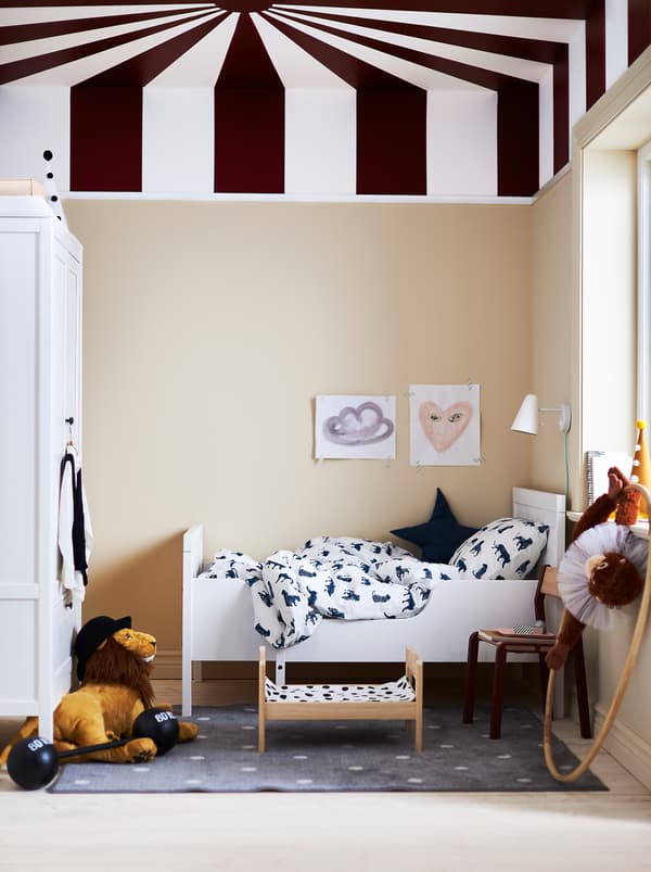 Um quarto de criança com uma cama extensível SUNDVIK, peluches e teto pintado com riscas radiantes, semelhante à cobertura de uma tenda de circo.