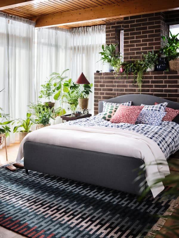 Um quarto luminoso com uma cama acolchoada HAUGA, um tapete RESENSTAD multicor e plantas verdes em todo o lado.