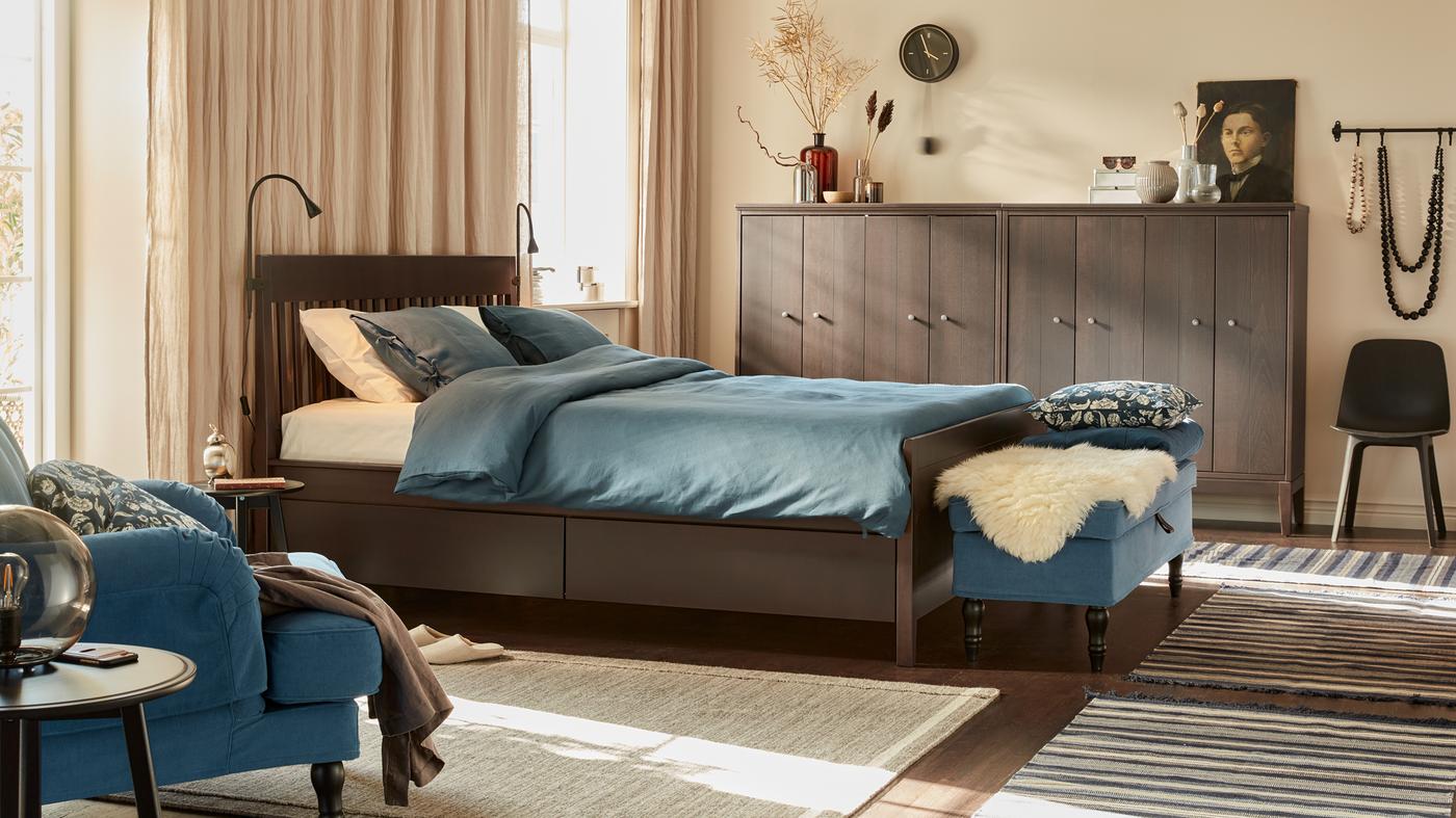 Um quarto tranquilo com paredes em bege, cortinados de linho, cama de madeira, têxteis em azul escuro, um banco e armários de madeira junto às paredes.