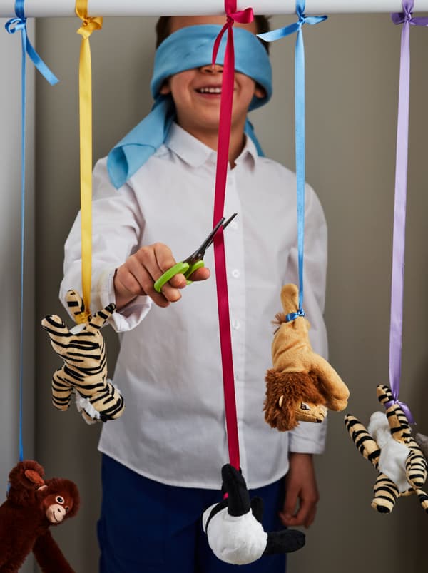 Um rapaz sorridente, com os olhos vendados, junto a uma fila de peluches DJUNGELSKOG pendurados em fitas, prestes a cortar uma delas com uma tesoura MÅLA.