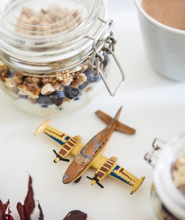 Um recipiente de vidro com granola e um avião de brincar sobre uma superfície branca.