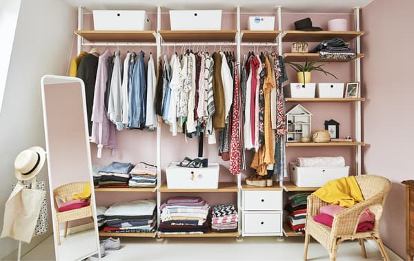 Um roupeiro aberto cheio de roupa com varões, prateleiras e caixas encostados a uma parede rosa.