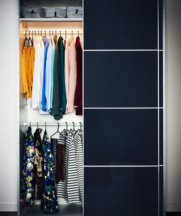 Um roupeiro com portas deslizantes com painéis abertas para um lado, a parte aberta apresenta duas filas de roupas muito ordenadas em cabides.