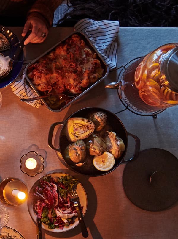 Um tacho em ferro fundido com tampa e ao lado um tabuleiro de forno com comida saborosa. As velas pequenas criam um ambiente aconchegante.