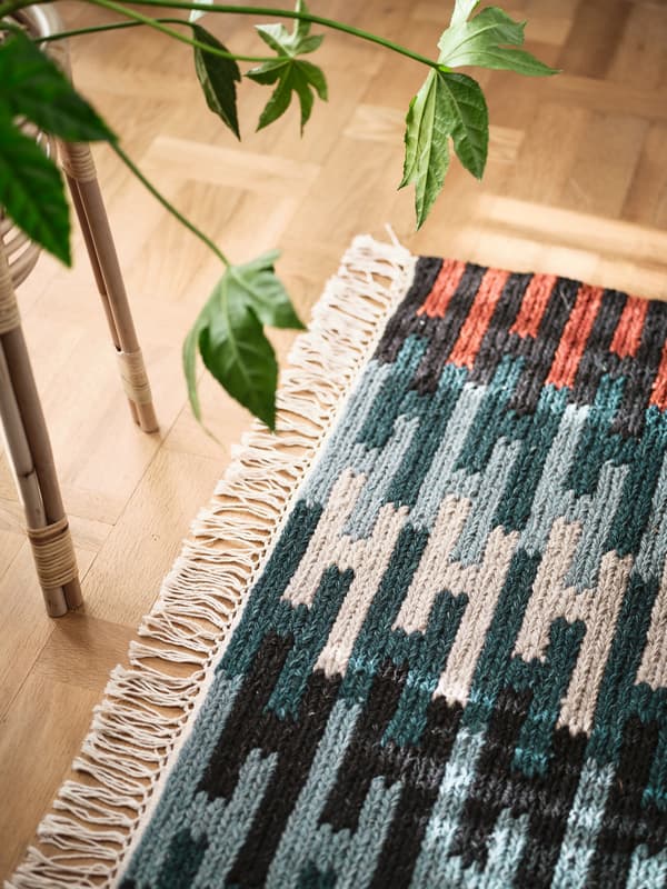 Um tapete RESENSTAD multicor está ao lado de uma mesa em bambu com uma planta verde em cima, numa divisão luminosa.