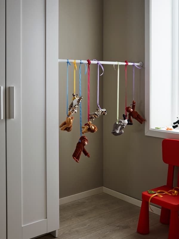 Um varão BOTAREN entre um roupeiro e uma parede no lado oposto, com uma fila de peluches DJUNGELSKOG pendurados em fitas coloridas.