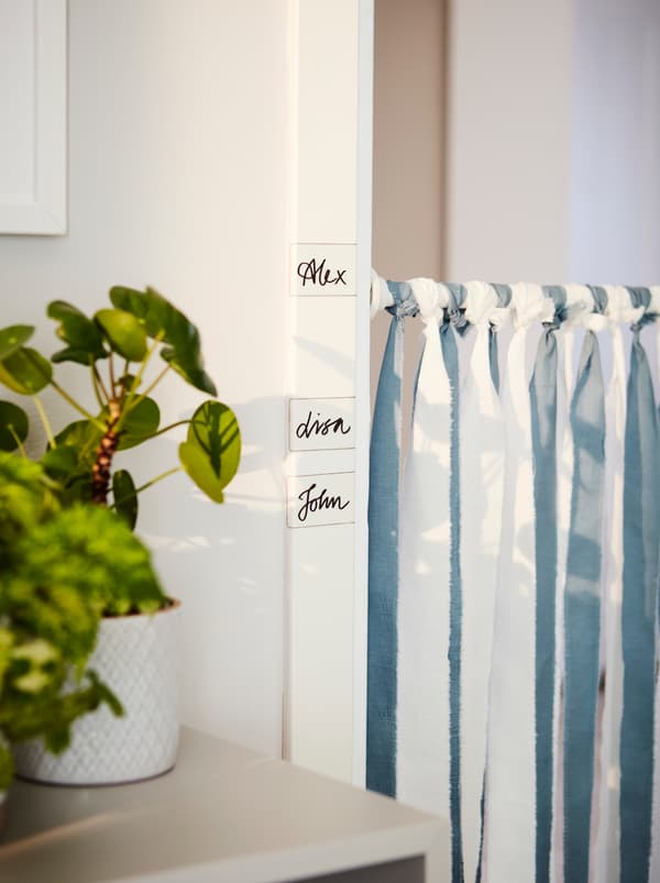 Um varão para cortina de duche BOTAREN está pendurado numa porta. Ao longo da ombreira da porta estão etiquetas IKEA 365+ com um nome escrito em cada.