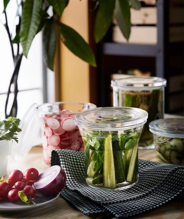 Uma bancada junto a uma janela. Em cima estão vários recipientes transparentes com tampa, cheios com rabanetes e pepinos em pickles.