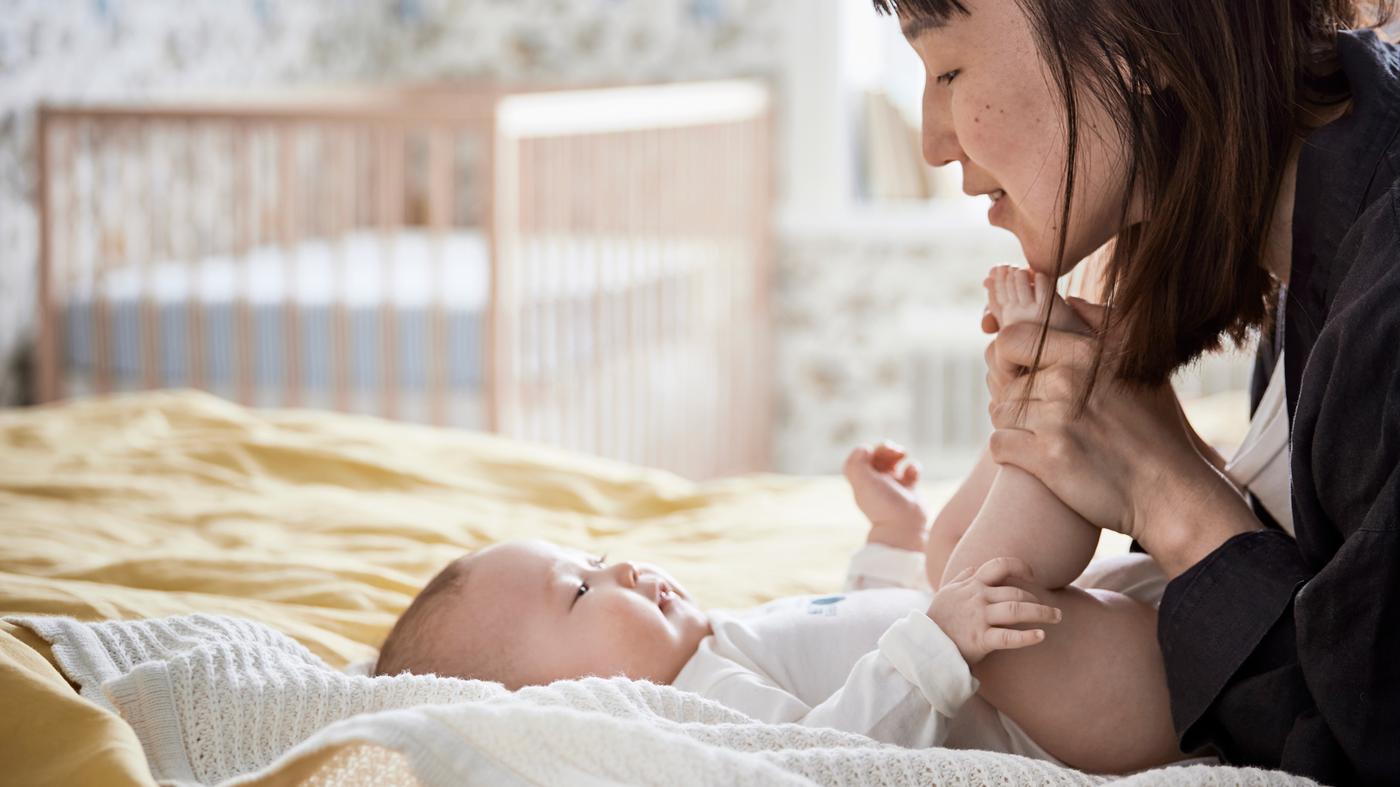 Uma bebé sorridente deitada sobre uma manta GULSPARV em cima de uma cama, com a mãe a segurar-lhe os pés e a sorrir para ela.