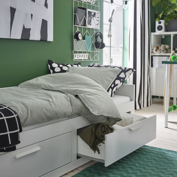Uma cama individual/dupla BRIMNES em branco está preparada para dormir. Uma gaveta está aberta e a roupa de cama e uma manta em verde estão guardadas no interior.