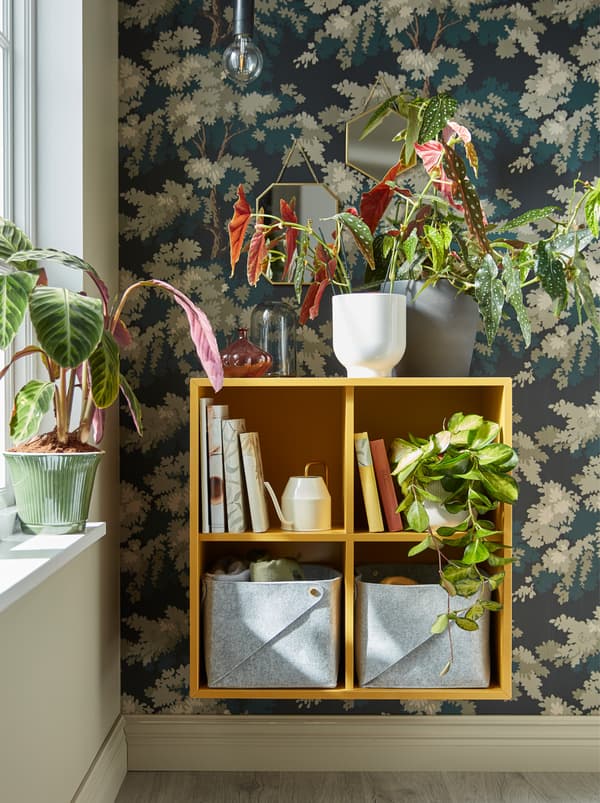 Uma estante EKET, em amarelo, montada na parede ao lado de uma janela, com plantas na parte de cima e livros no interior.