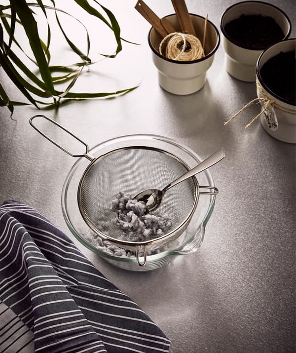 Uma grande taça de vidro com um passador IDEALISK que contém pasta de papel húmida, feita de guardanapos cortados em pedaços, ao lado de três vasos para plantas.