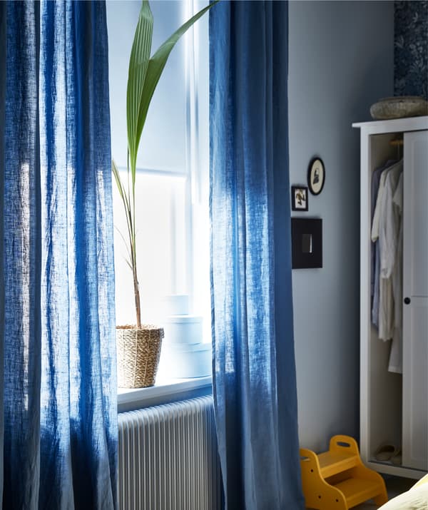 Uma janela com painéis brancos, cortinados transparentes em azul e um vaso, ao lado de um roupeiro aberto.