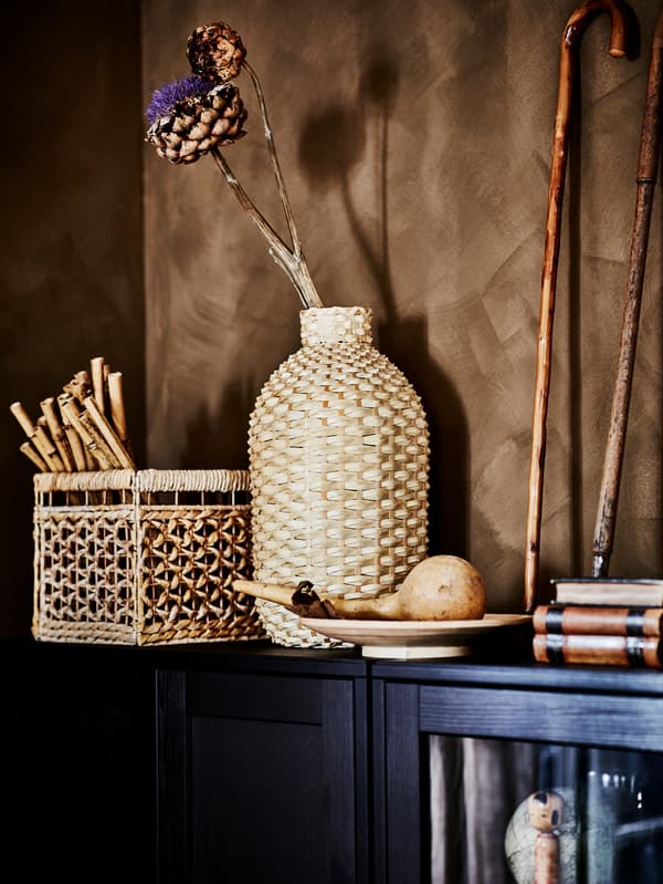 Uma jarra KAFFEBÖNA em bambu com uma flor de alcachofra seca no interior, entre outros objetos artísticos no topo de dois armários HAVSTA de cor escura.