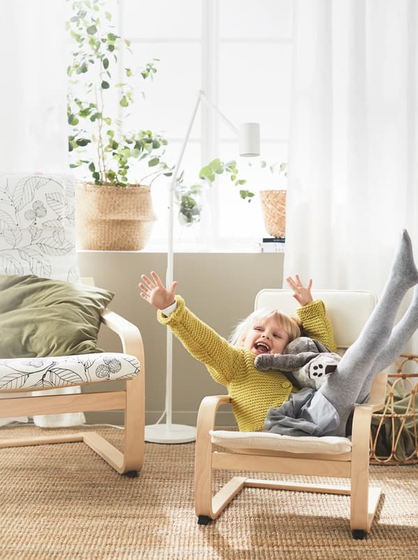 Uma menina alegremente sentada de pernas e braços abertos, com um peluche, numa poltrona para criança POÄNG, junto a uma poltrona de tamanho normal.