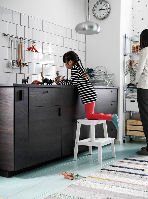 Uma menina, de joelhos em cima de um escadote com 2 degraus BEKVÄM, brinca com pequenos animais de brincar em cima de uma bancada de cozinha escura. Uma mulher está perto.