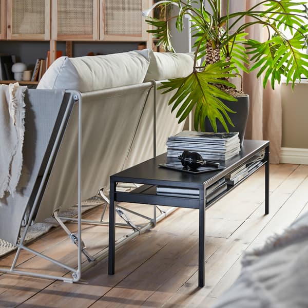 Uma mesa de centro comprida, esguia, em preto, colocada ao longo da parte de trás de um sofá. Em cima da mesa está uma pilha de revistas e uma planta grande.