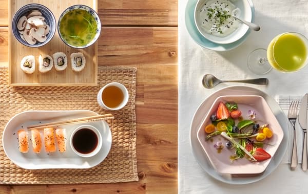 Uma mesa posta de duas maneiras: uma para sushi num prato branco oval, outra para uma salada num prato rosa hexagonal.