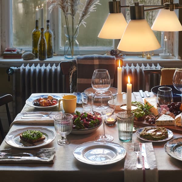 Uma mesa pronta para a refeição com uma toalha de uma só cor, panos de cozinha a servir de guardanapos e uma mistura informal de pratos e copos.