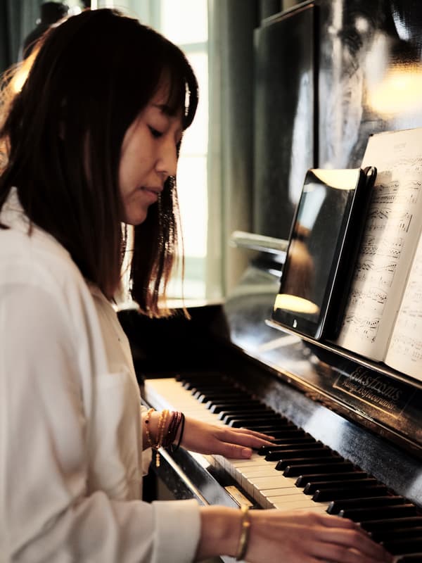 Uma mulher com cabelo comprido escuro junto a um piano, com as mãos nas teclas.