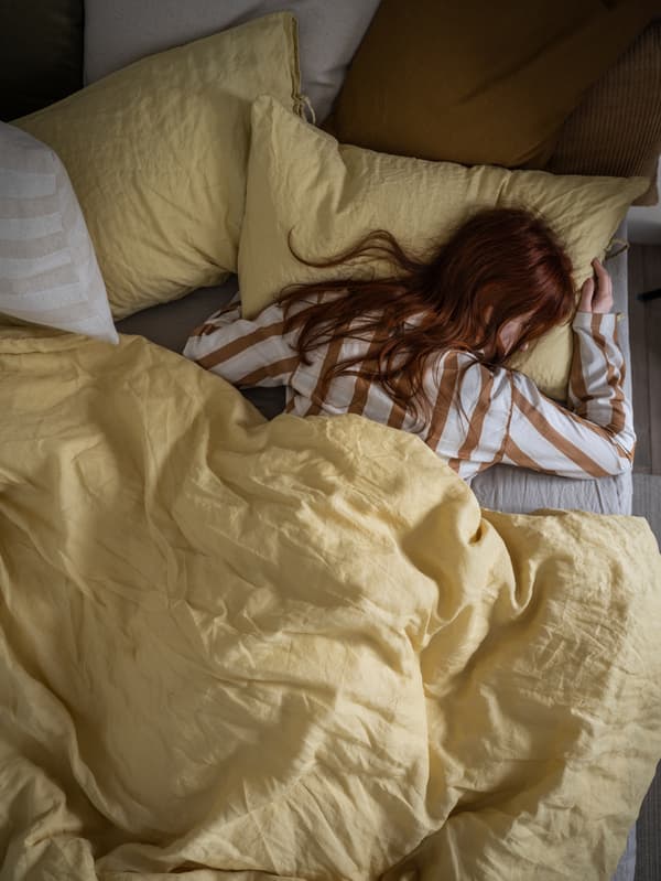 Uma mulher com um pijama às riscas a dormir profundamente numa cama com uma capa de edredão e fronhas PUDERVIVA, em amarelo claro.