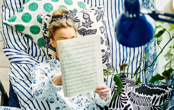 Uma mulher deitada na cama com almofadas, roupa de cama e papel de parede à sua volta com diferentes padrões, enquanto lê pautas musicais.