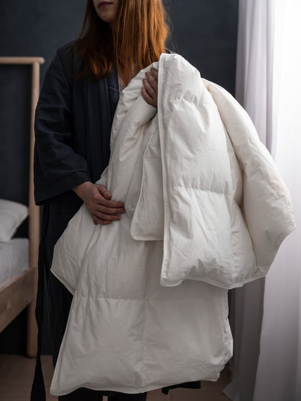 Uma mulher junto a uma cama num quarto com pouca luz, com paredes cinzentas com cortinados transparentes, em branco, a segurar num edredão FJÄLLARNIKA.
