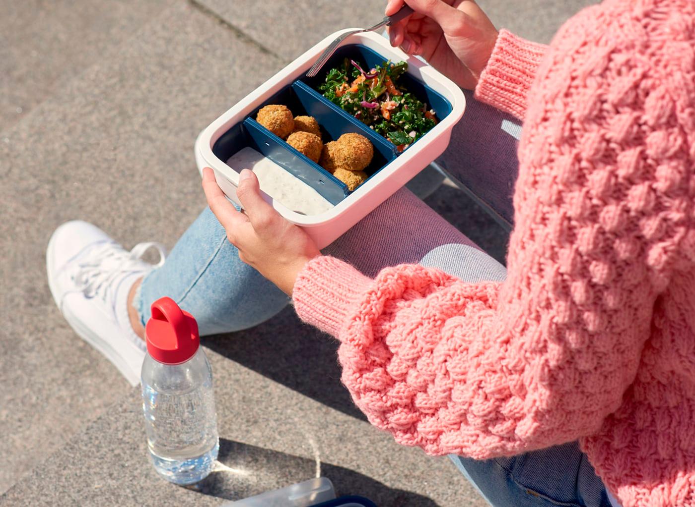Uma mulher sentada a comer de um recipiente para alimentos em plástico, com uma garrafa de água em plástico transparente ao seu lado.