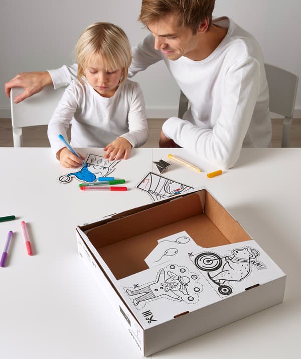 Un adulte assis avec un enfant en train de colorier des images de cirque, découpées dans la boîte en carton sur la table.