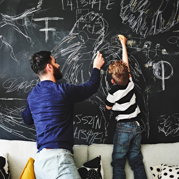 Un adulte et un enfant agenouillé et debout sur un canapé, en train de dessiner à la craie sur un mur. Des coussins sont éparpillés contre le dossier du canapé.