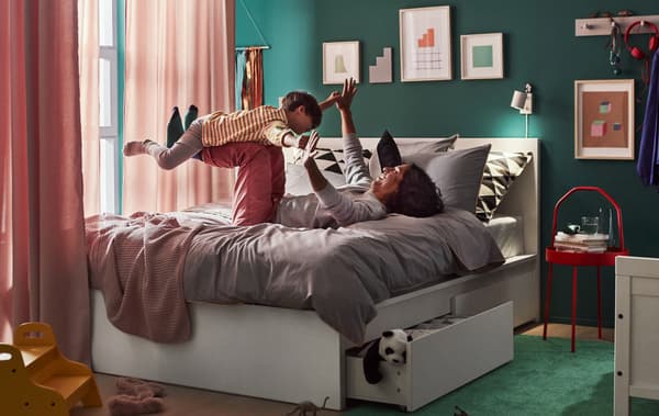Un adulte et un enfant jouant sur un lit double avec tiroirs de rangement sous le lit, avec des rideaux montés autour du lit et des œuvres d’art aux murs.