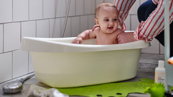 Un adulto con una camicia a righe bianche e rosse regge un bambino in una vaschetta LÄTTSAM. Accanto alla vaschetta, un tappetino per vasca da bagno PATRULL verde - IKEA.