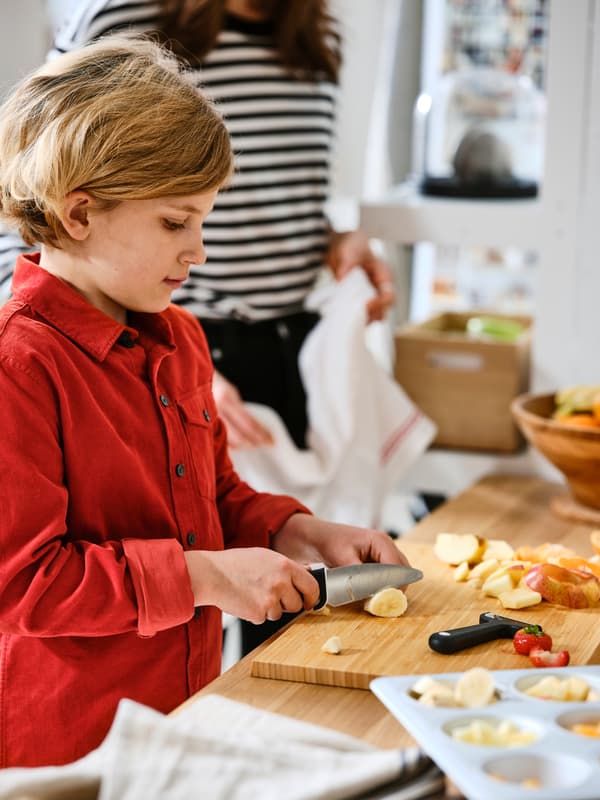 Un adulto guarda un bambino con una camicia rossa intento a tagliare frutta su un tagliere APTITLIG in bambù servendosi di un coltello e un utensile per sbucciare SMÅBIT - IKEA.