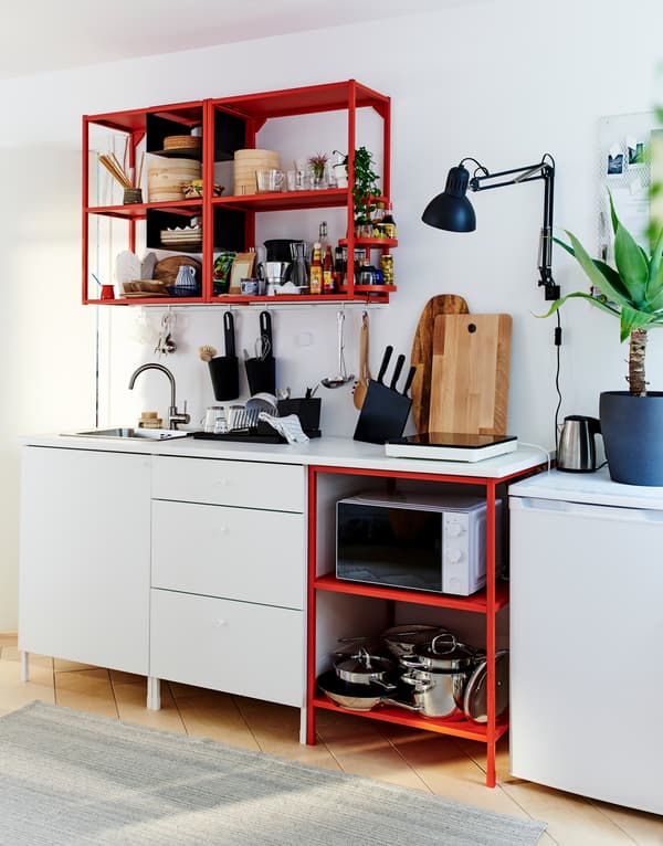 Un agencement de cuisine élémentaire avec des tiroirs blancs, des tablettes rouges en métal pour le rangement au sol et sur le mur, et un petit réfrigérateur.