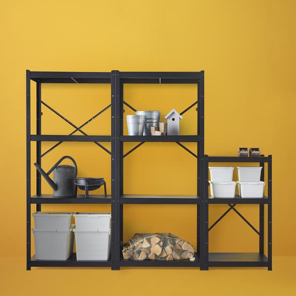 Un agencement d’étagères BROR noires contre un mur orange clair, avec du bois de chauffage, des boîtes de rangement blanches et des outils de jardinage.