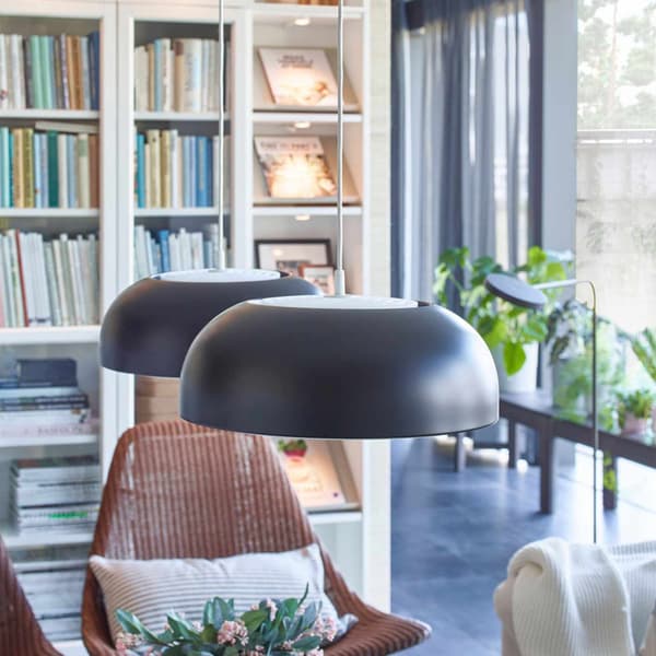 Un angolo di un soggiorno con in fondo una libreria bianca con diversi libri al suo interno e delle piante vicino alle finestre. In primo piano due lampadari appesi al soffitto due sedie in vimini rosse. 