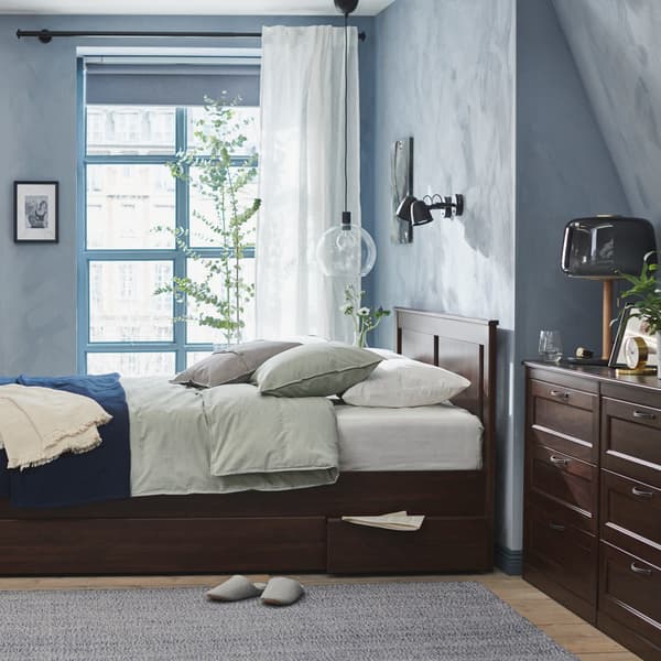 Un angolo di una camera da letto con un letto, un tappeto GÅNGVÄG su cui sono appoggiate delle ciabatte. All'angolo della stanza è posizionata una cassettiera marrone SONGESAND. 