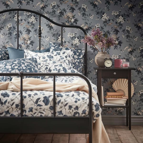 Un angolo di una stanza con una struttura letto SAGSTUA e con affianco un comodino con sopra dei fiori, una sveglia e altri oggetti. 