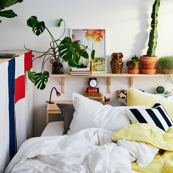 Un angolo di una stanza da letto. Sul letto un copripiumino bianco ÄNGSLILJA. Dietro il letto due mensole con sopra una sveglia, dei libri e delle piante. 