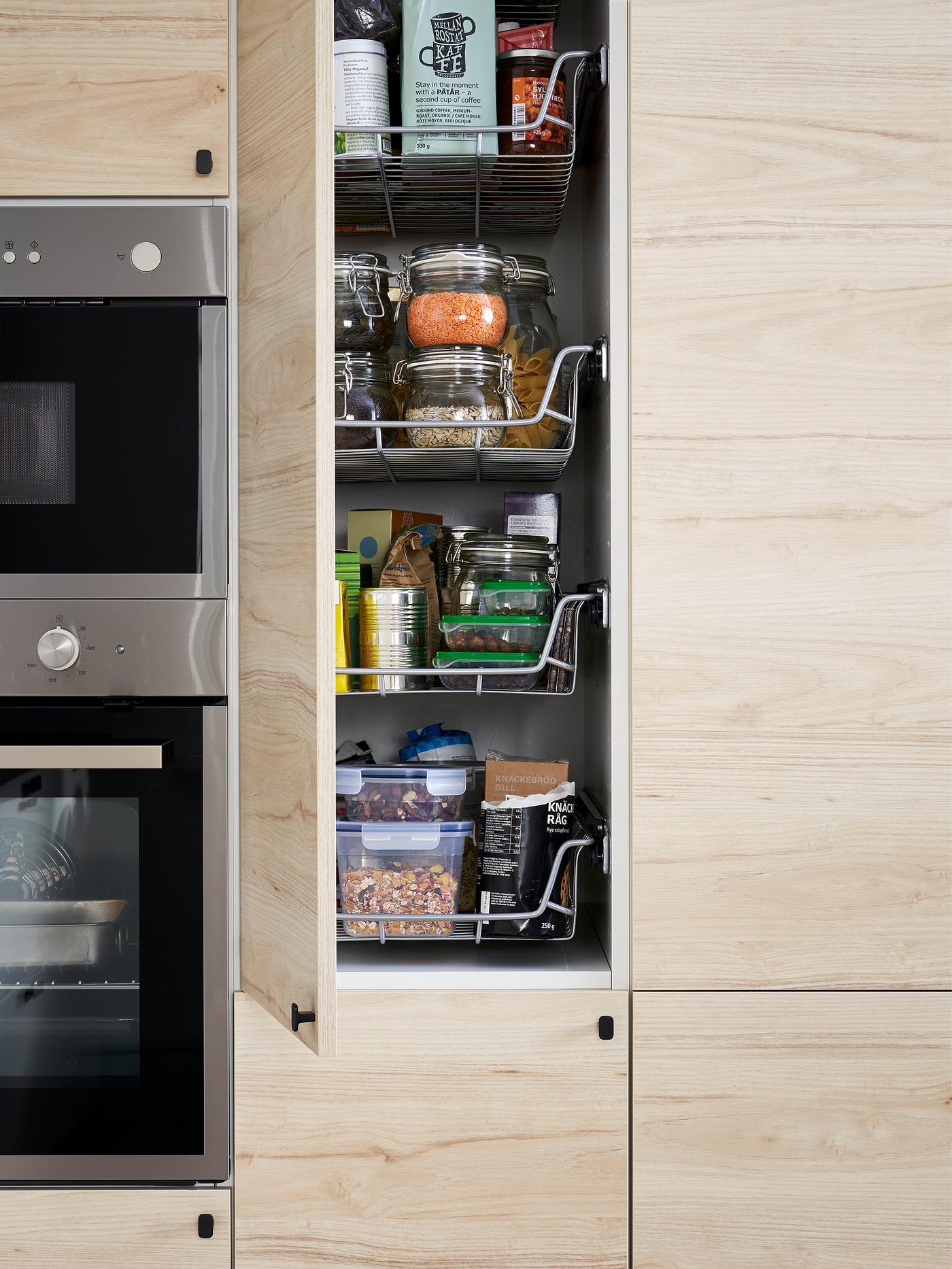 Un armario alto con una puerta abierta de efecto fresno claro dentro de una cocina. Dentro hay cestos de rejilla, recipientes de alimentos y otros objetos.