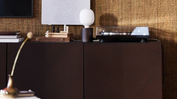 Un armario BESTÅ en marrón oscuro contra una pared de arpillera, una lámpara de mesa BLEKKLINT y un marco de fotos con altavoz wifi SYMFONISK.