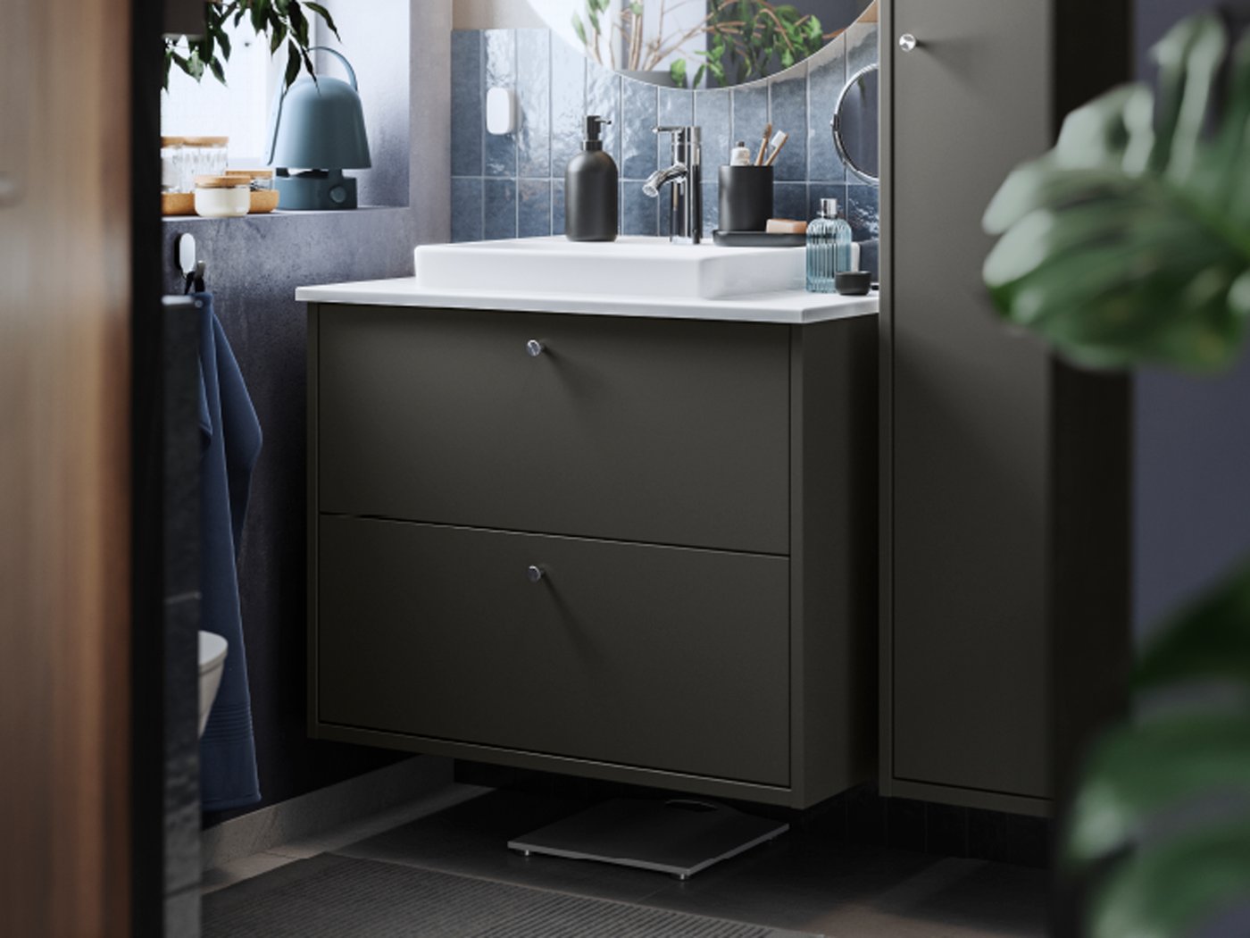 Un armario de lavabo HAVBÄCK en gris escuro con caixóns e un lavabo; hai un armario de parede alto en gris escuro ao lado e un espello enriba.